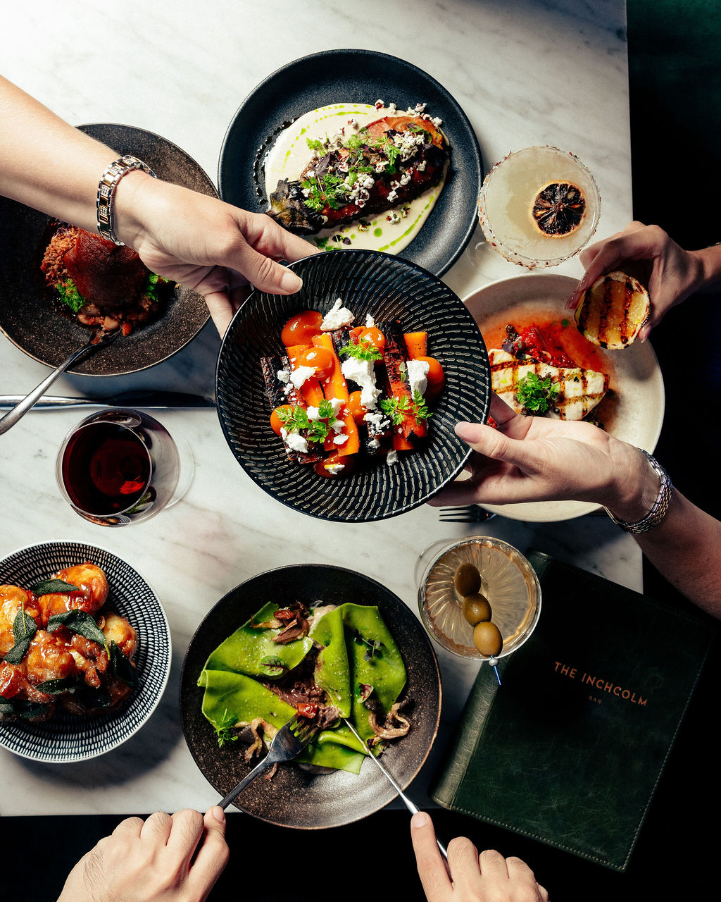 Overhead view of hands sharing a table of plated dishes, cocktails, and wine.