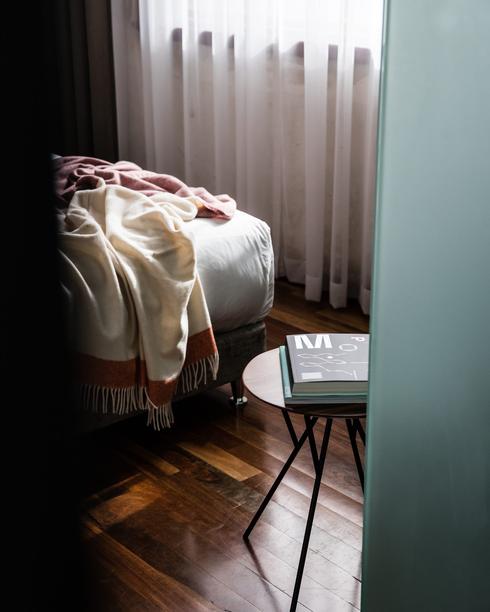 A sofa draped with blankets sits beside a bright curtained window, with books stacked on a small coffee table.