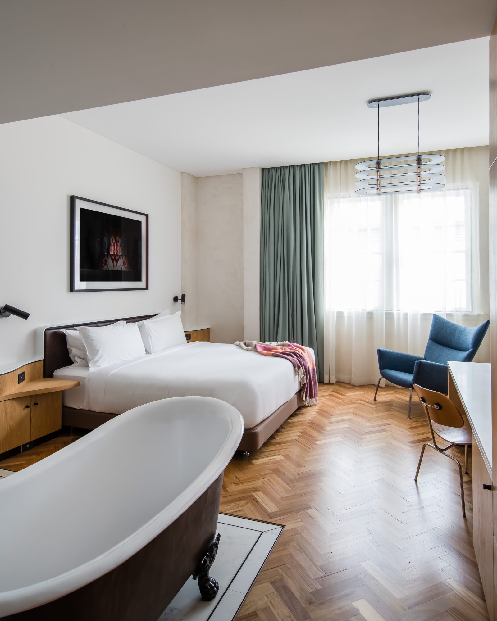 Modern bedroom with a white bed, freestanding bathtub, wood flooring, teal curtains, and a sculptural pendant light.