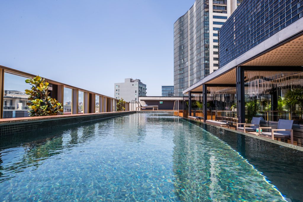 Rooftop swimming pool bordered by lounge chairs and greenery, with modern high-rise buildings under a clear blue sky.