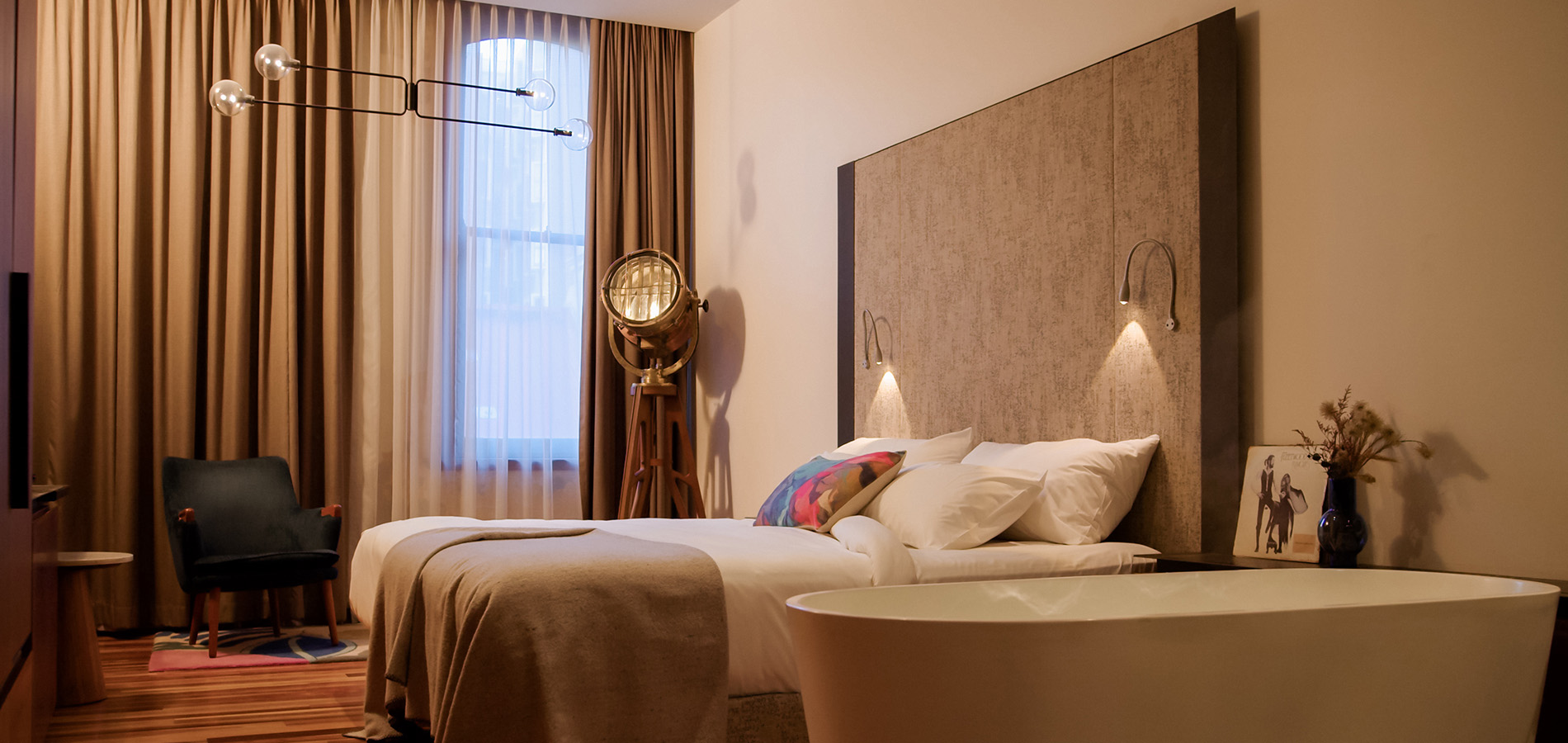 Modern bedroom with a white bed, freestanding bathtub, curtained window, dark armchair, and warm wall lights.