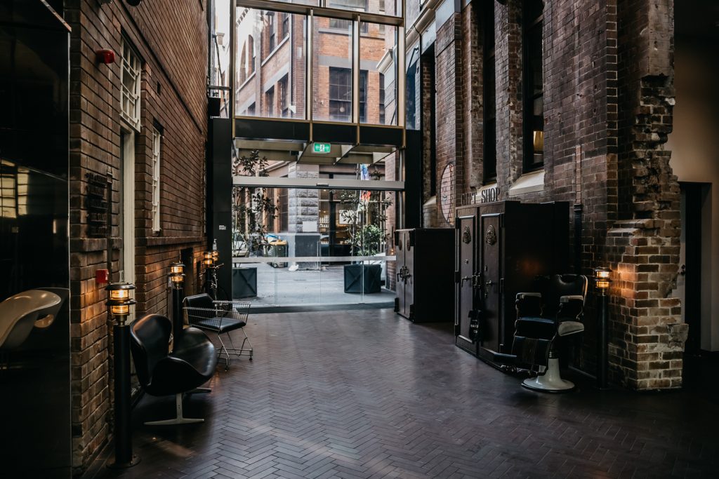 Brick-lined interior passage with vintage barber chairs, a City Shop sign, and glass doors opening to a courtyard.