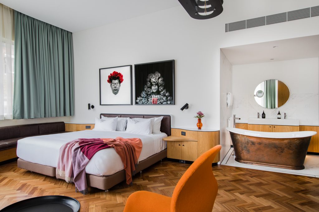 Modern hotel bedroom with a white bed, colorful blankets, teal curtains, framed portraits, and a freestanding copper bathtub.