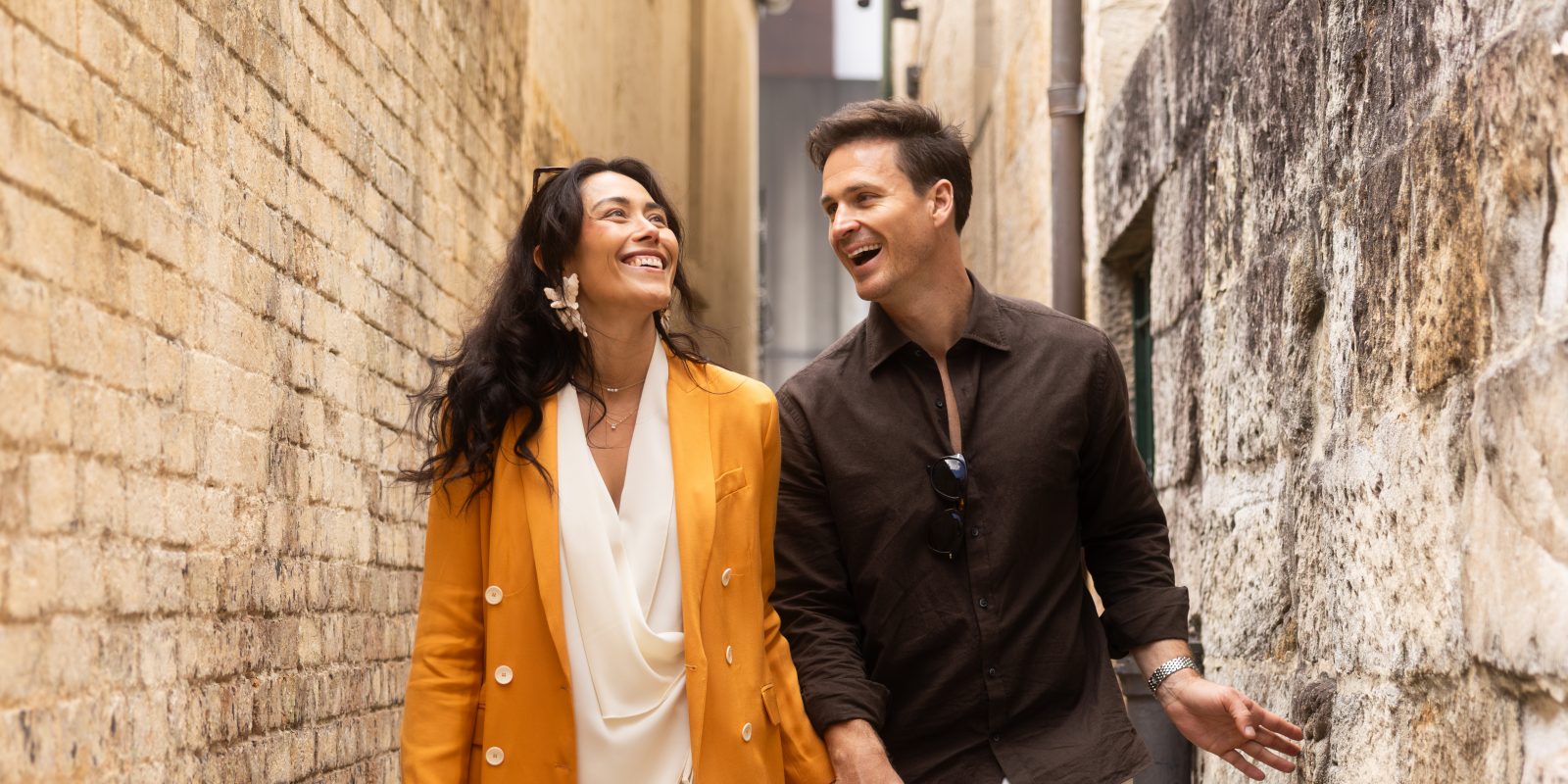 A smiling couple stands arm in arm beside a weathered stone wall; she wears a mustard blazer, and he wears a dark shirt.