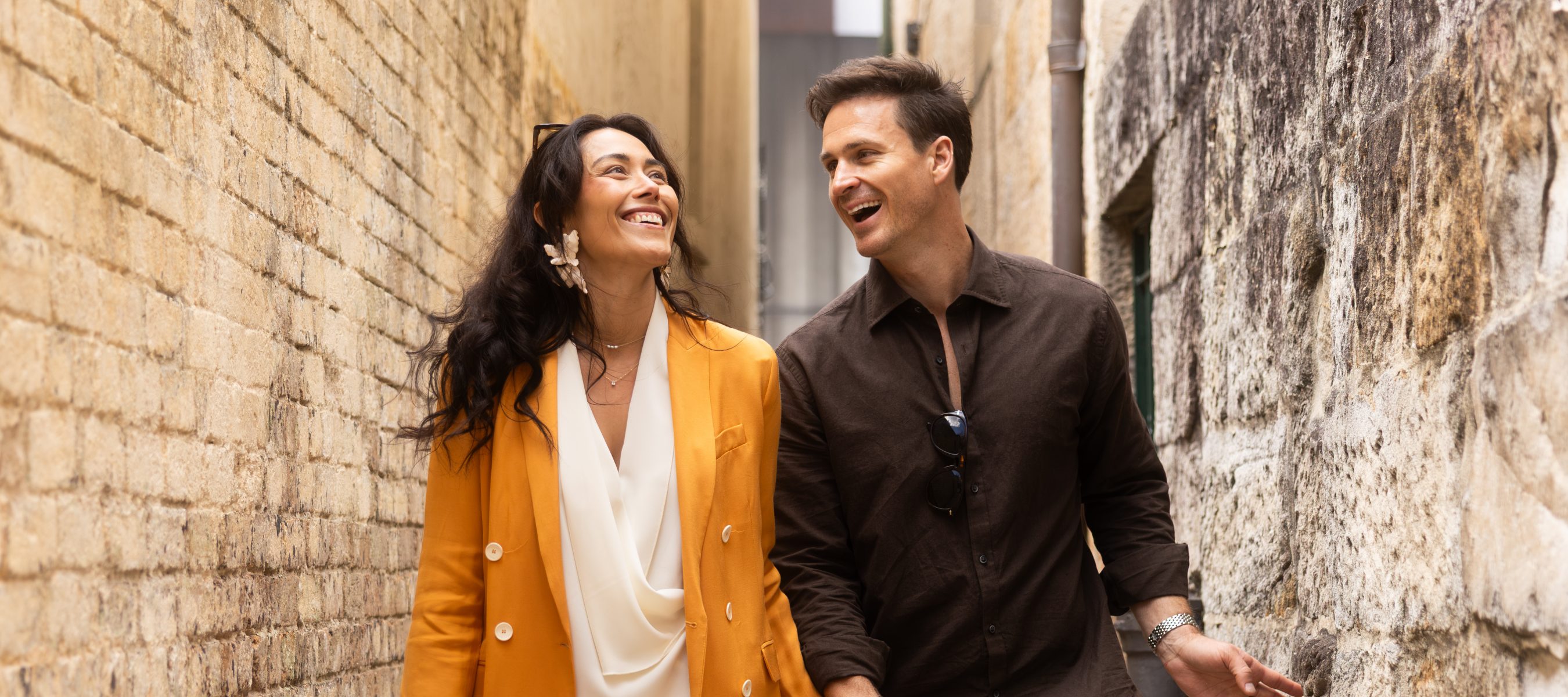 A smiling couple stands arm in arm beside a weathered stone wall; she wears a mustard blazer, and he wears a dark shirt.