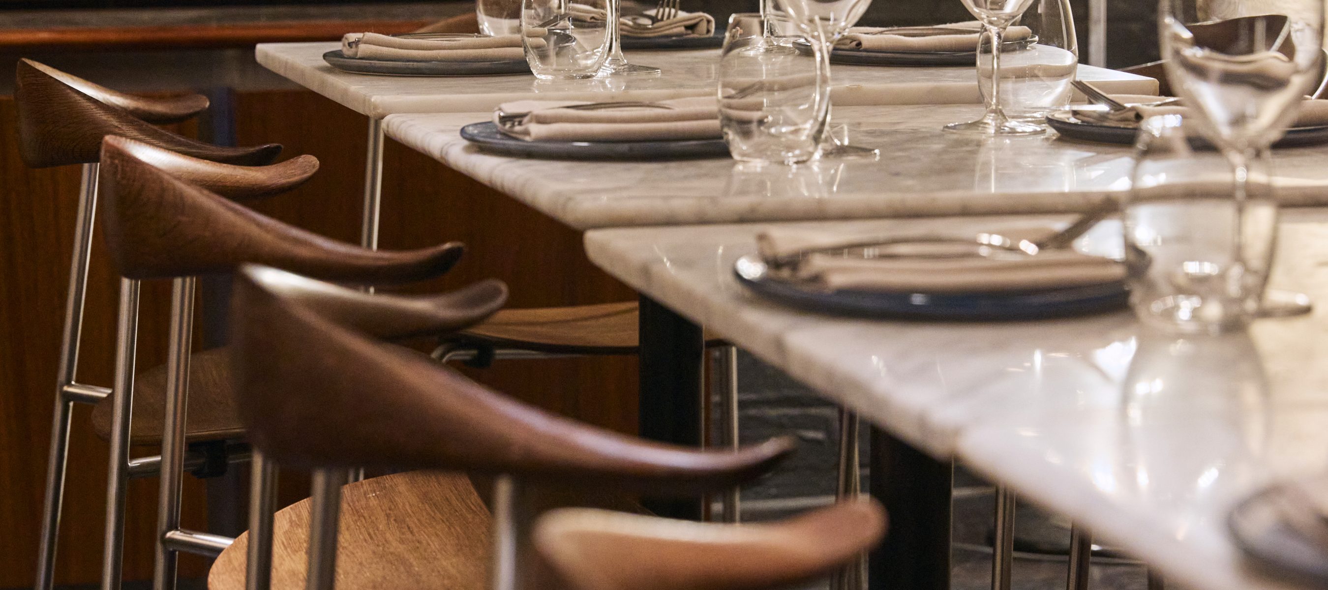 A row of marble dining tables set with plates, folded napkins, and wine glasses, surrounded by wooden chairs.