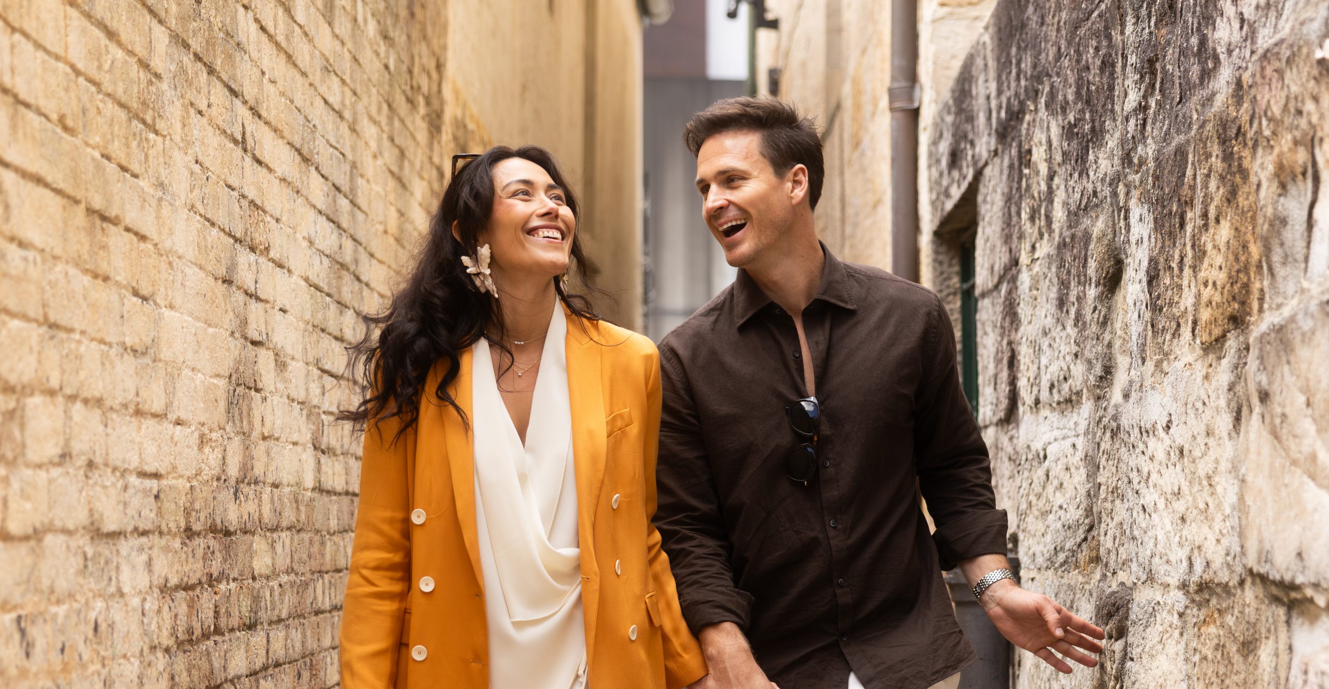 A smiling couple stands arm in arm beside an old stone wall, with a narrow street behind them.